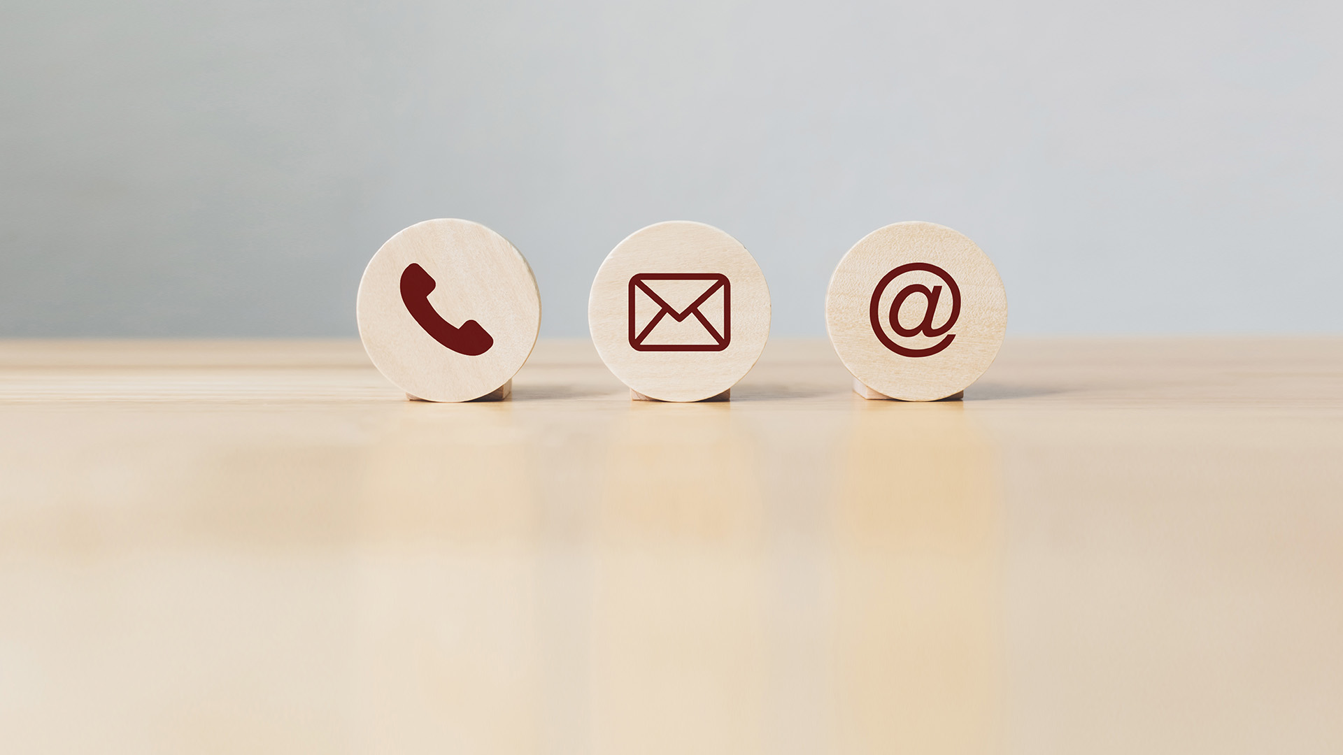 The image displays three small, square wooden signs on a light-colored surface with a blurred background. Each sign has a different symbol  the first has an envelope icon, the second is a telephone handset, and the third depicts a computer mouse cursor.