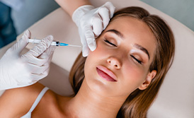 A woman receiving a cosmetic treatment, with a professional applying a product to her face using a wand-like tool.